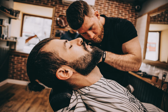 Getting perfect shape. Close-up side view of young stylish bearded man getting beard haircut by bearded, muscular hairdresser or barber at barbershop - Powered by Adobe