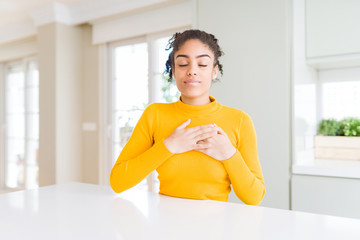 Beautiful african american woman with afro hair wearing a casual yellow sweater smiling with hands on chest with closed eyes and grateful gesture on face. Health concept.