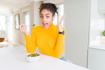 Young african american woman eating healthy green vegatables very happy and excited, winner expression celebrating victory screaming with big smile and raised hands