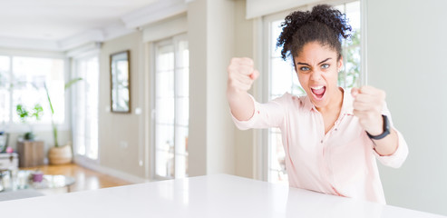 Wide angle of beautiful african american woman with afro hair angry and mad raising fists frustrated and furious while shouting with anger. Rage and aggressive concept.
