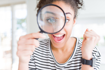 Young african american woman looking through magnifying glass screaming proud and celebrating...