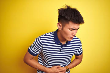 Young asian chinese man wearing striped polo standing over isolated yellow background with hand on stomach because indigestion, painful illness feeling unwell. Ache concept.