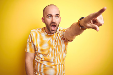 Young bald man with beard wearing casual striped t-shirt over yellow isolated background Pointing with finger surprised ahead, open mouth amazed expression, something on the front