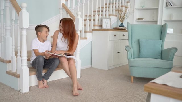 son and mom sits on the stairs and read a book . They look at camera and smile