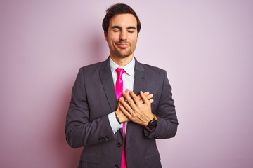 Young handsome businessman wearing suit and tie standing over isolated pink background smiling with hands on chest with closed eyes and grateful gesture on face. Health concept.