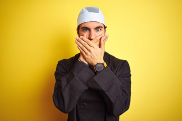 Young handsome chef man cooking wearing uniform and hat over isolated yellow background shocked covering mouth with hands for mistake. Secret concept.