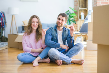 Young beautiful couple moving to a new house sitting on the floor with a big smile on face, pointing with hand and finger to the side looking at the camera.