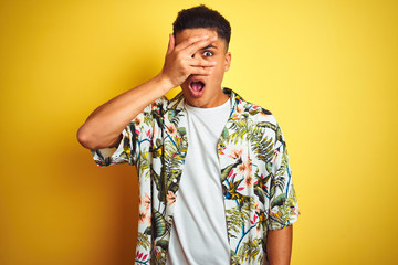 Young brazilian man on vacation wearing summer floral shirt over isolated yellow background peeking in shock covering face and eyes with hand, looking through fingers with embarrassed expression.