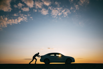 Silhouette of man driver pushing his car along on an empty road after breakdown at sunset, copy space, side view. Road trip, trouble on the road  - obrazy, fototapety, plakaty