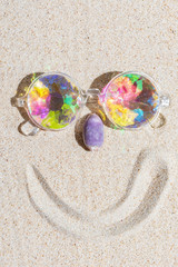 Fototapeta na wymiar Funny smiling face made of kaleidoscope multicolor glasses and purple sea shell on sand background with copy space, flat lay, vertical composition