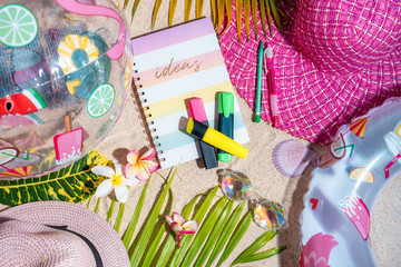Colourful writing note pad with pink and green pen on sand, surrounded by green palm leafs, sea shells, pink hat, inflatable toys. Summer beach background, flat lay