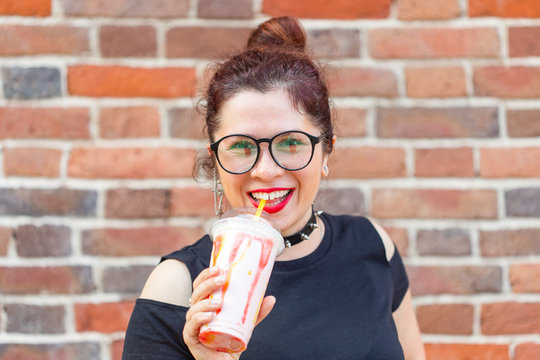 Young positive girl holds in Ruach a milkshake with a straw against a brick wall background. Summer dessert and weekend break concept.