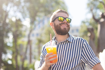 Portrait of a positive cheerful young man with a glass of juice with a straw while walking in the park on a warm sunny summer day. The concept of rest after study and work on weekends.