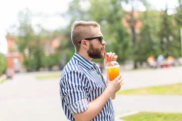 Fototapeta premium Portrait of a positive cheerful young man with a glass of juice with a straw while walking in the park on a warm sunny summer day. The concept of rest after study and work on weekends.