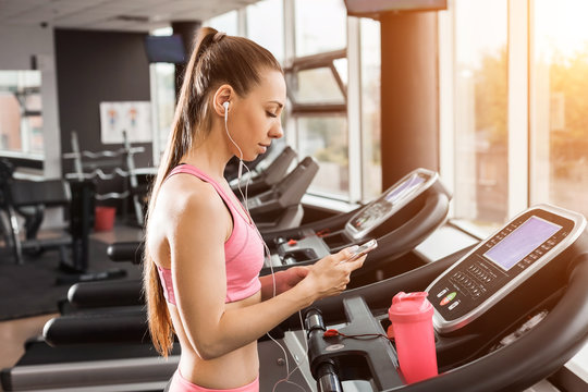 slim beautiful girl with a ponytale standing on the running track in the gym holding a phone wearing earphones listening to music. Concept of cardio exercises and healthy way of life