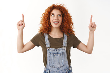 Impressed curious young redhead stylish curly-haired woman, looking up amused and entertained, smiling enthusiastic white toothy grin, pointing gazing top copy space promo, standing white background