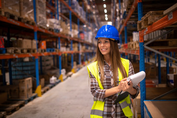 Portrait of cheerful woman in protective uniform checking packages and stock of products in warehouse storage room. Industrial worker controlling distribution in storehouse.