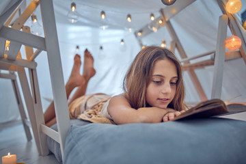 Little 10 year old girl reading classic book under her home-made tent inside the living room.