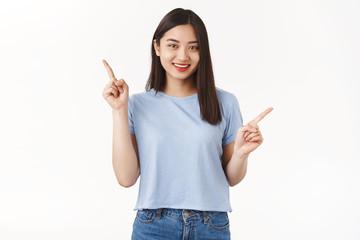 Cute attractive happy cheerful brunette short-haired asian girl pointing sideways two variants left upper promo smiling joyfully carefree picking products have choices white background