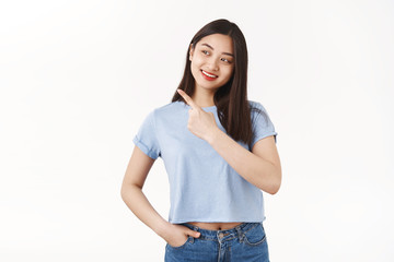 Interested curious pretty asian brunette girl student intrigued looking pointing left side copy space tilt head amused happy broad smile pick right good choice standing white background