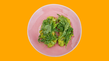 Two slices of mashed avocado toast garnished with fresh rocket or arugula salad set on a pink plate against a bright orange background, ideal snack or meal for a vegetarian or a plant-based diet
