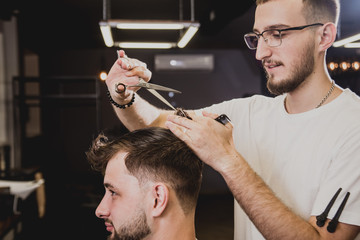 Fototapeta premium Young man with trendy haircut at barber shop. Barber does the hairstyle and beard trim.