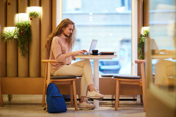 Full length side view portrait of beautiful teenage girl using laptop in cafe while studying for college, copy space