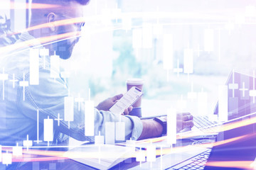Candlestick chart in foreground. In background man sits, uses smartphone, works on laptop. Businessman reads message in phone.