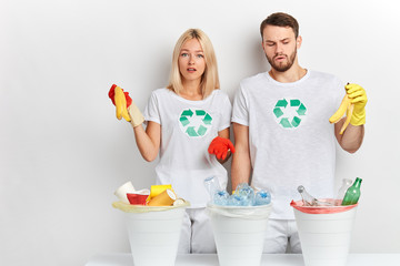 serious woman and handsome man standing near three trash cans, holding banana peel, being puzzled where to throw them family sorting rubbish, throwing banana peel and looking at camera