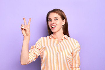 Front view of funny cute girl showing peace gesture, has great joyful mood, showing white teeth, smiling wide, dressed in comfortable casual shirt with stripes, posing over light purple wall.