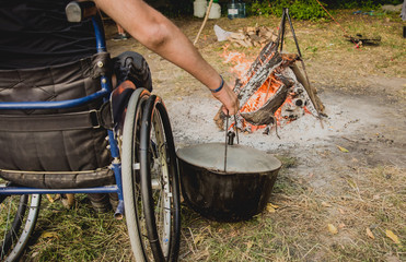Disabled man resting in a campsite with friends. Wheelchair in the forest on the background of bonfire. Barbeque. Camping.