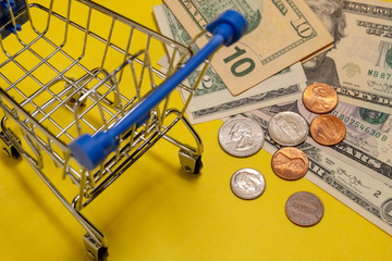 Close-up of US dollars and cents next to a shopping trolley in a supermarket. Yellow background. Concept of shopping, discounts, online shopping, consumer ability or black friday.