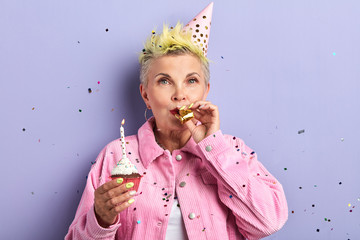 stylish woman in party hat blowing noisemaker, celebrating birthday, holiday party. isolated blue...