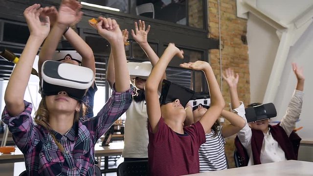 Group of six schoolchildren wearing virtual reality headset with raised armsworking on new programing project during class