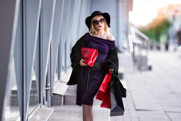 Stylish blonde woman in black coat, sunglasses and hat with shopping bags walks along the street