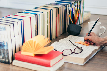 Stack of books education background, female hand makes notes near open textbook. Glasses, pens and pencils in holder, cup of tea with lemon.