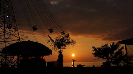 Fototapeta premium Swinging carousel roundabout chain ride at sunset. Entertainment on the beach, silhouettes of palm trees on a background of sea sunset