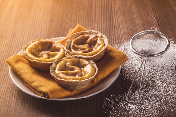 Obraz na płótnie Canvas Delicious handmade artisan Belem cakes on a napkin on a plate. Small strainer with icing sugar to decorate the cream cakes on a rustic wooden table. Sunlight enters from the side. Portuguese pastry.