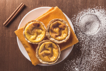 Top view of delicious handmade artisan Belem cakes on a napkin on a plate. Small strainer with icing sugar to decorate the cream cakes on a rustic wooden table. Cinnamon stick. Portuguese pastry.