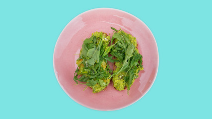 Two slices of mashed avocado toast garnished with fresh rocket or arugula salad set on a pink plate against an aqua blue background, ideal snack, lunch or dinner for a vegetarian or a plant-based diet