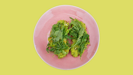Two slices of mashed avocado toast garnished with fresh rocket or arugula salad set on a pink plate against a lime green background, ideal snack, lunch or dinner for a vegetarian or a plant-based diet