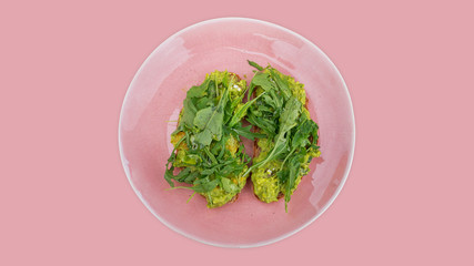 Two slices of mashed avocado toast garnished with fresh rocket or arugula salad set on a pink plate against a pink background, ideal snack, lunch or dinner for a vegetarian or a plant-based diet