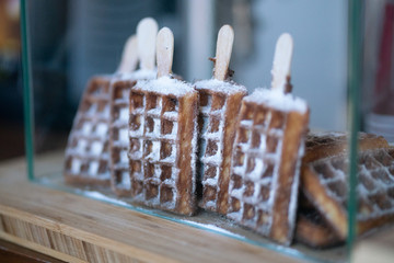 Freshly baked belgium waffles on a wooden stick, sprinkled with powdered sugar. delicious dessert behind the glass in the store.