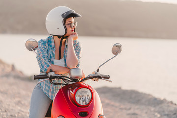 Young pretty woman's portrait. Girl in white helmet and eyeglasses sitting on red retro scooter at nature background.