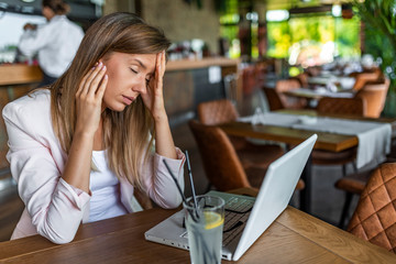 Portrait of tired young business woman with laptop computer. Tired businesswoman having a headache while working on her laptop in cafe shop. Beautiful tired business woman is touching her forehead