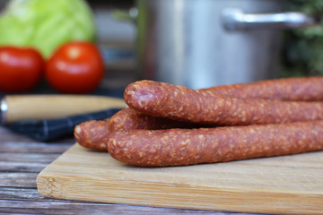 Red German ham sausages on wooden plate arranged with knife and vegetables in blurry background