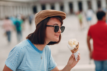 Young smiling  woman with cone gelato ice cream in hand in striped frock vest,  white pants and cap at  Piazza San Marco place, Venice, Veneto, Italy