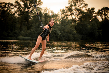 Girl riding on the wakeboard on the river in the sunset holding a rope of the motorboat