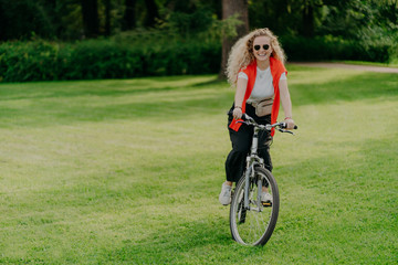 Outdoor image of pretty cheerful young woman rides bike, wears sunglasses, casual wear, poses on green lawn, spends free time in park, bikes in beautiful nature. Activity and recreation concept