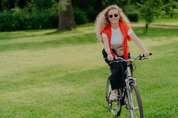 Fototapeta na wymiar People, nature, rest, lifestyle concept. Happy curly woman rides bicycle among green grass, moves actively, wants to be fit, explores new places in countryside, wears sunglasses, casual clothes